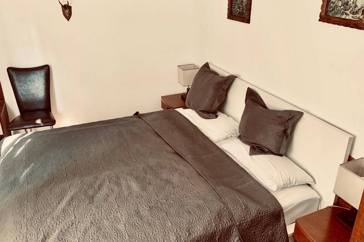Een net opgemaakt bed met een bedspreide met patroon, een stoel, nachtkastjes en decoraties aan de muur.