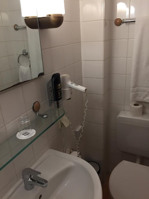 A bathroom with a glass shelf holding a glass, a hand dryer, and a toilet paper holder.