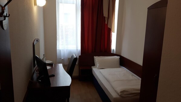 Una habitación de hotel pequeña con una cama individual, un escritorio con silla, una lámpara y una ventana con cortinas.