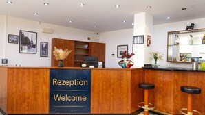 Una recepción con un letrero que dice 'Reception Welcome', un mostrador de madera, un estante con objetos decorativos, un espejo y una zona de bar con taburetes.