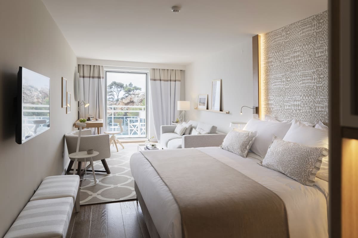 Une chambre d’hôtel moderne dotée d’un grand lit, d’un coin salon avec une table et des chaises, d’un balcon avec vue et d’un panneau décoratif mural.