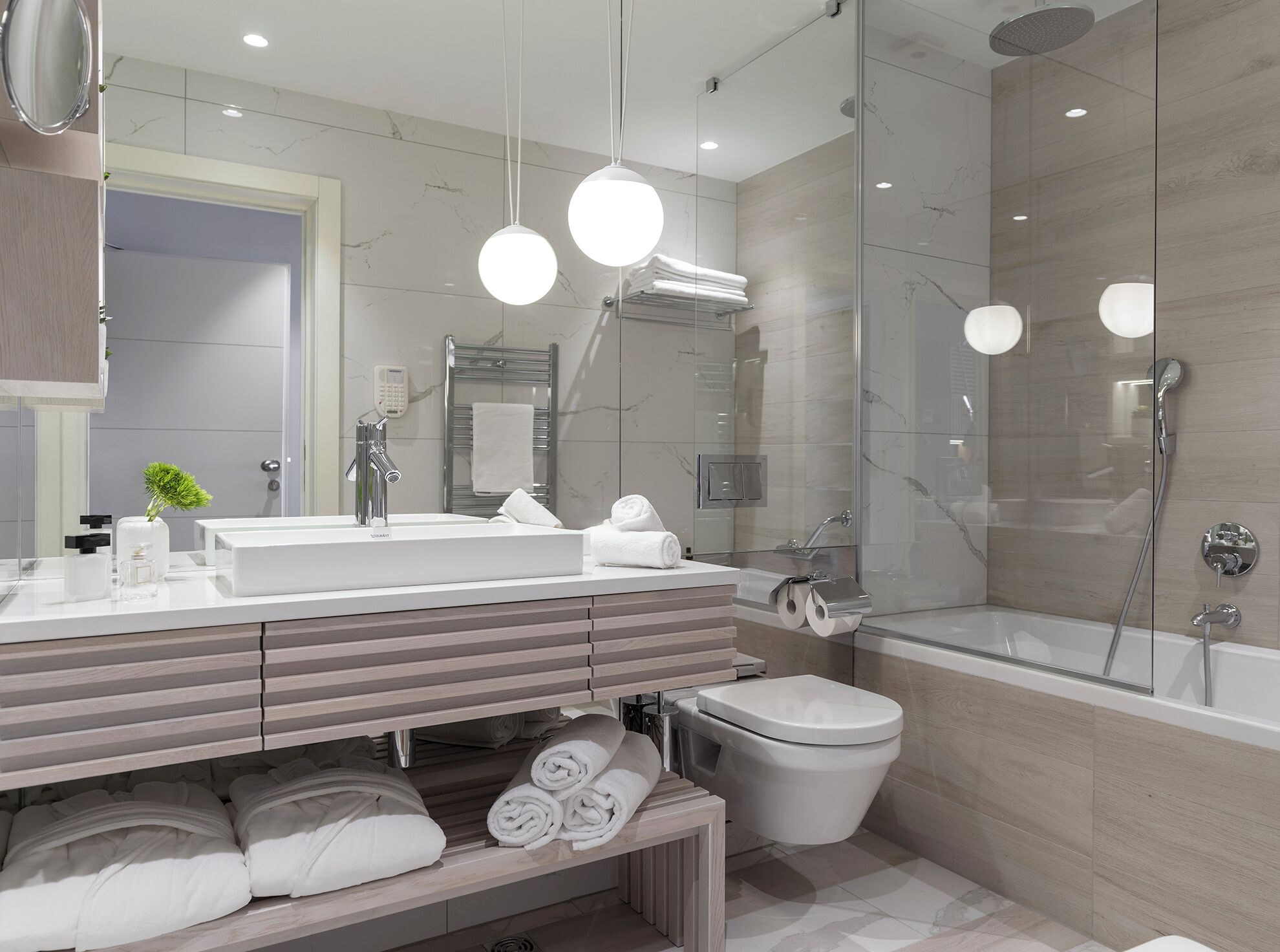 Un baño moderno con un espejo grande, un lavamanos, un inodoro y una ducha. Hay toallas y artículos de aseo en la encimera.