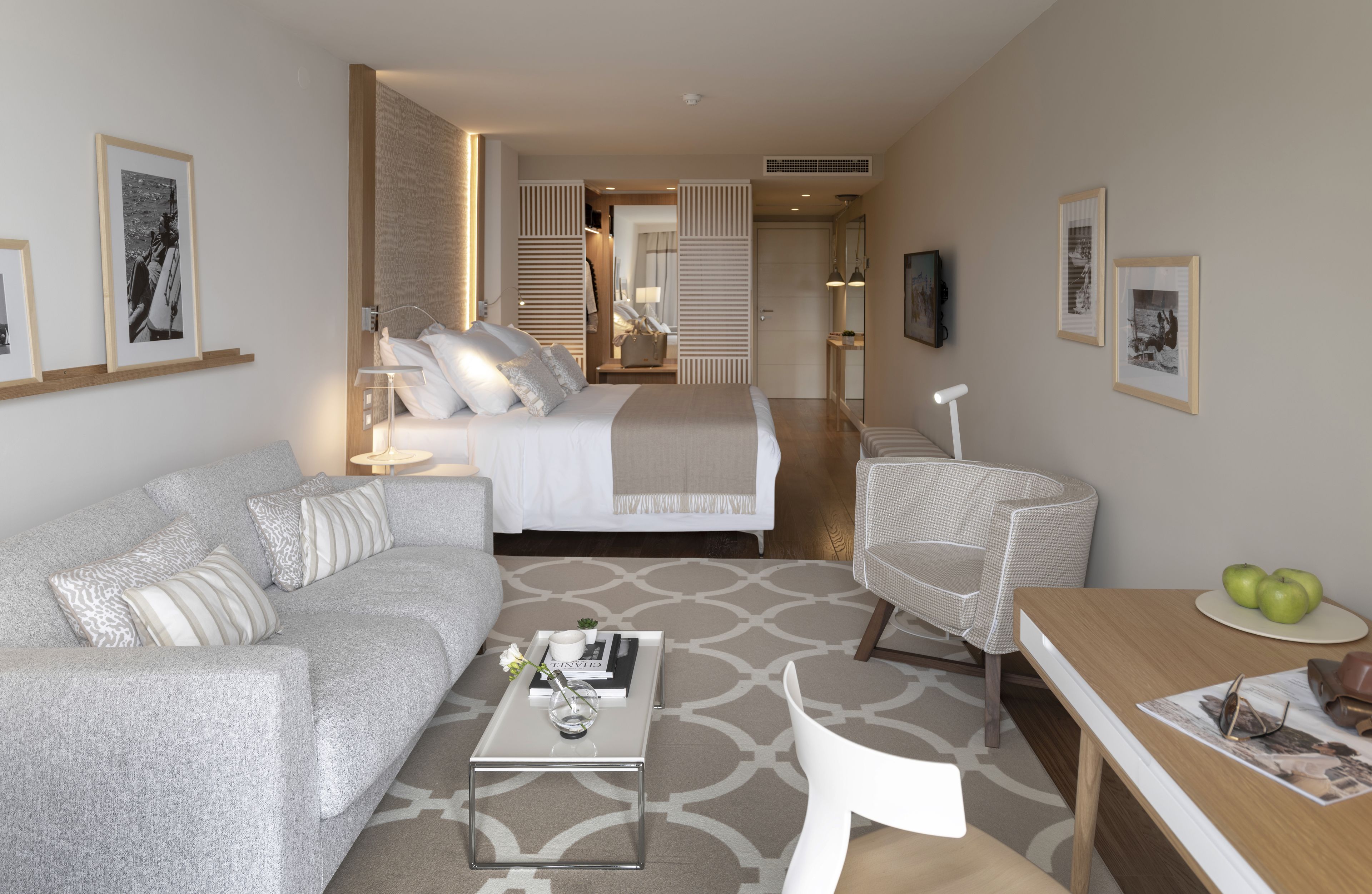 Quarto de hotel moderno com cama, sofá, mesa de jantar e cadeira. Há quadros nas paredes e uma pequena mesa com um vaso e um livro.