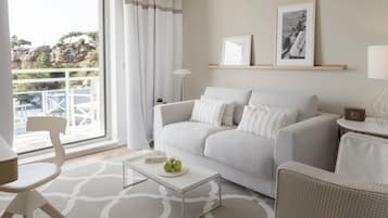 Un salon moderne avec un canapé gris, des coussins blancs, une table basse avec des pommes, une chaise blanche et une vue sur un balcon doté d’une balustrade.