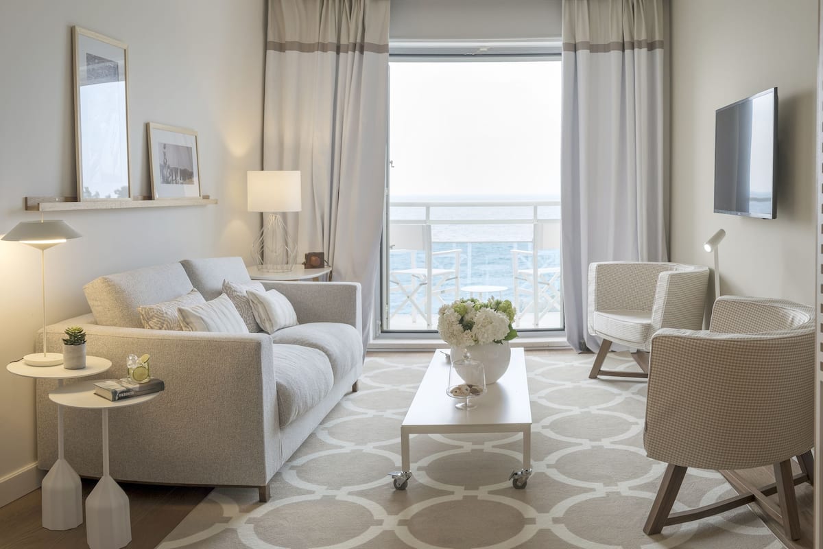 Une chambre d’hôtel moderne équipée d’un canapé, de deux fauteuils, d’une table basse, d’une télévision et d’un balcon avec des rideaux.