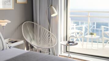 A modern hotel room with a large window, a white chair, a small round table with a book and glasses, and a lamp on a bedside table.