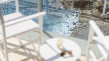 Guarda-corpo de vidro com vista para uma piscina, onde a água está a espirrar, um toldo e uma mesa pequena com uma garrafa.