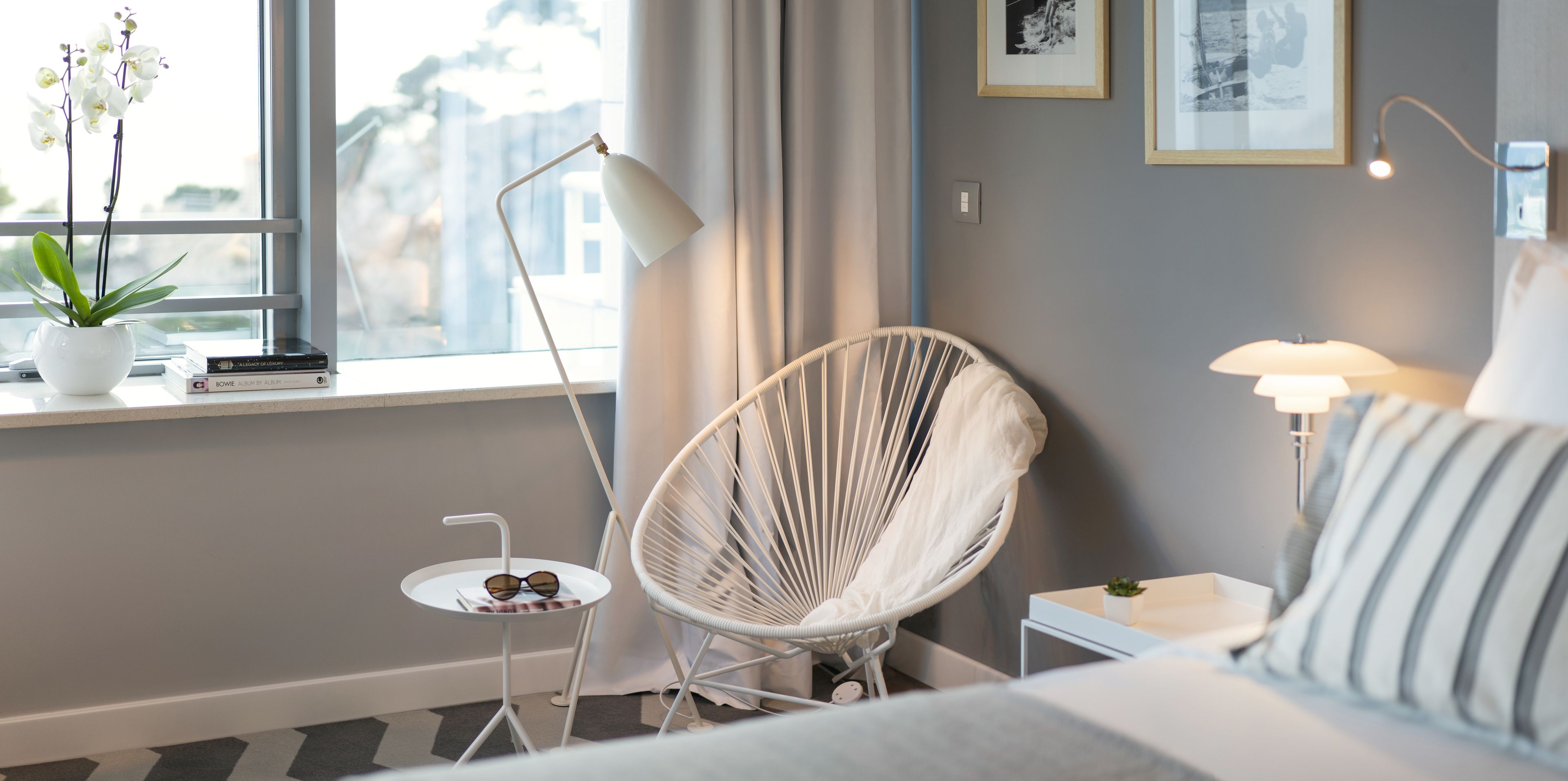 Una habitación de hotel moderna con una cama, una silla blanca, una mesita con una planta, una lámpara y una ventana con cortinas.