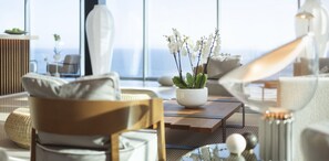A modern living room with a large window, a white vase with white flowers, a wooden coffee table, and a sofa with cushions.