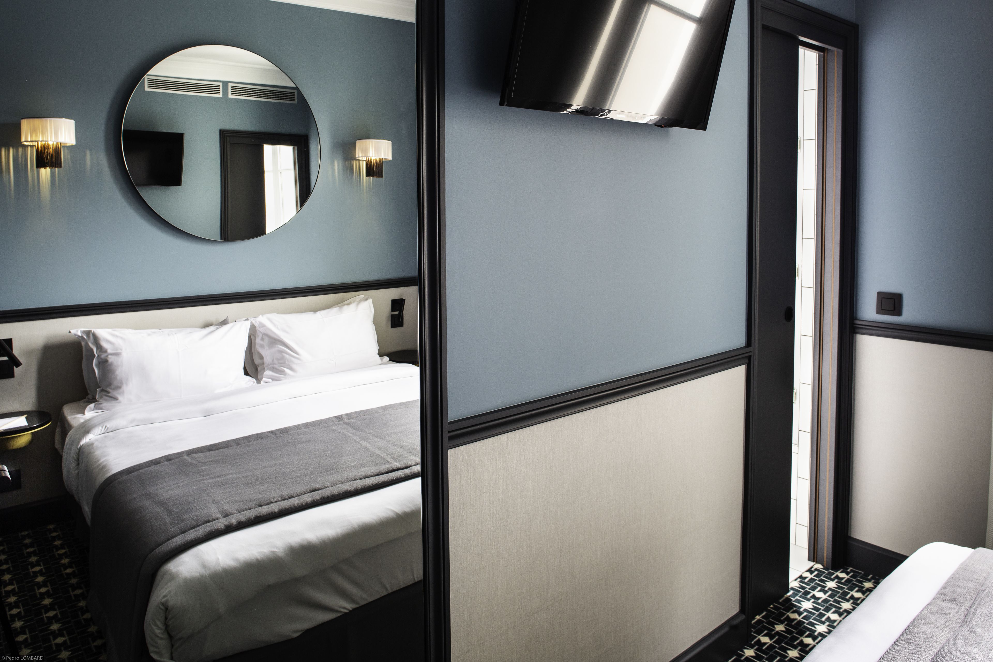A hotel room with a bed, a round mirror, a flat-screen TV, and a wall-mounted light fixture.