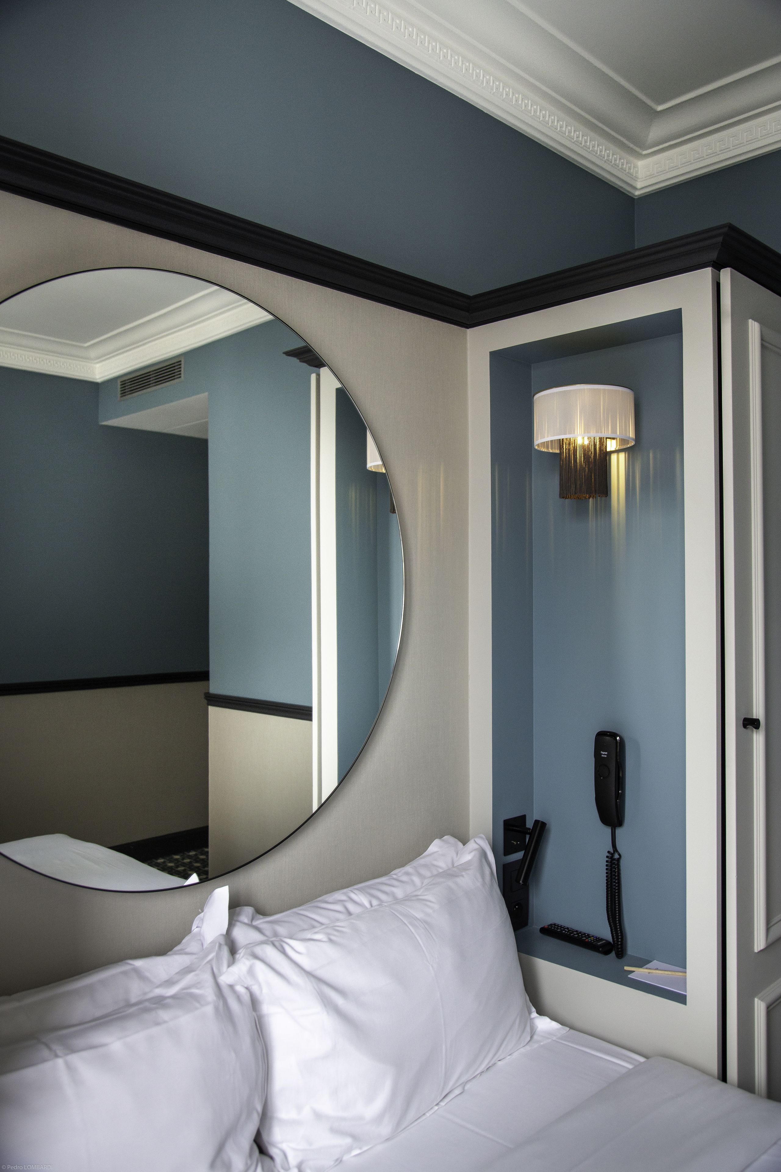 A hotel room with a large mirror, a bed with white linens, a bedside telephone, and a wall-mounted lamp.