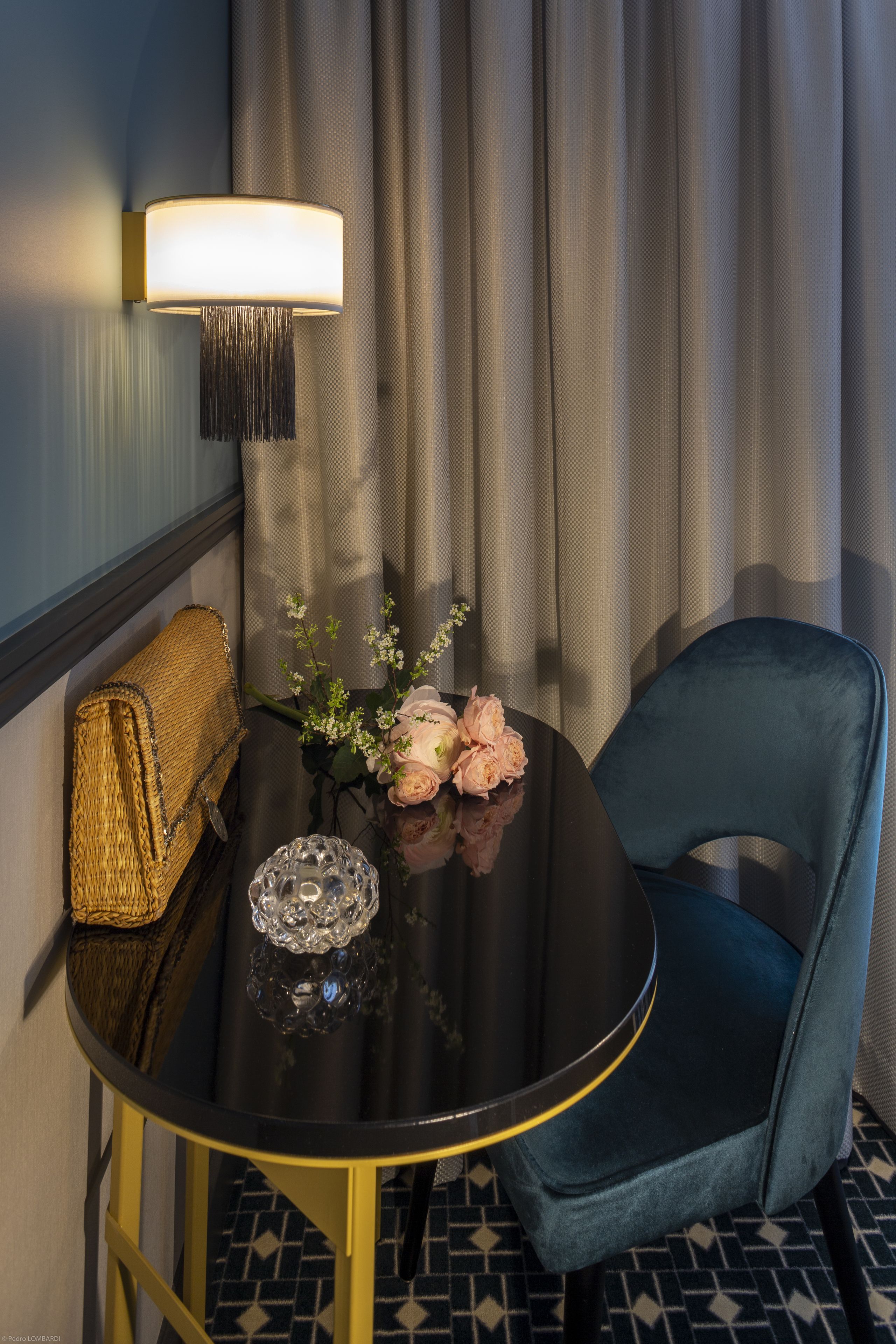A room with a round black table, a blue chair, a lamp, a woven basket, and a vase with flowers.