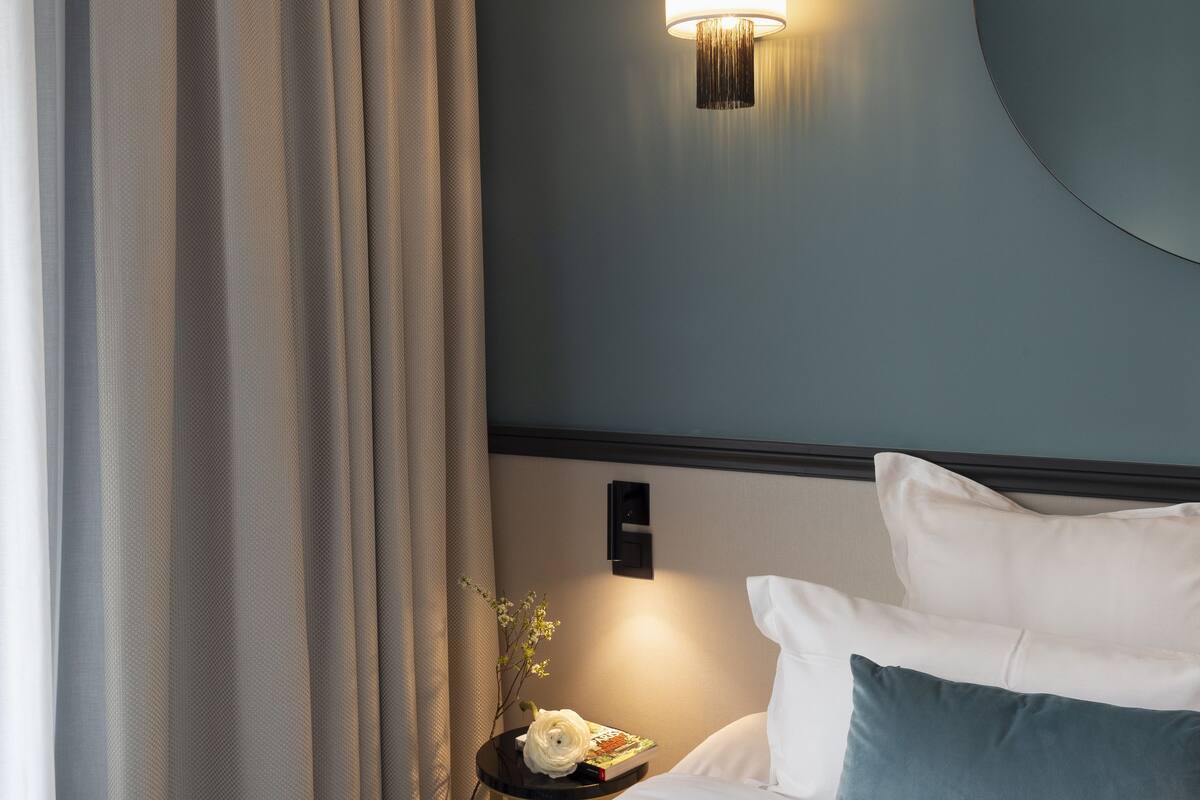 A neatly made bed with white and blue bedding, a round mirror on the wall, and a lamp on a bedside table.