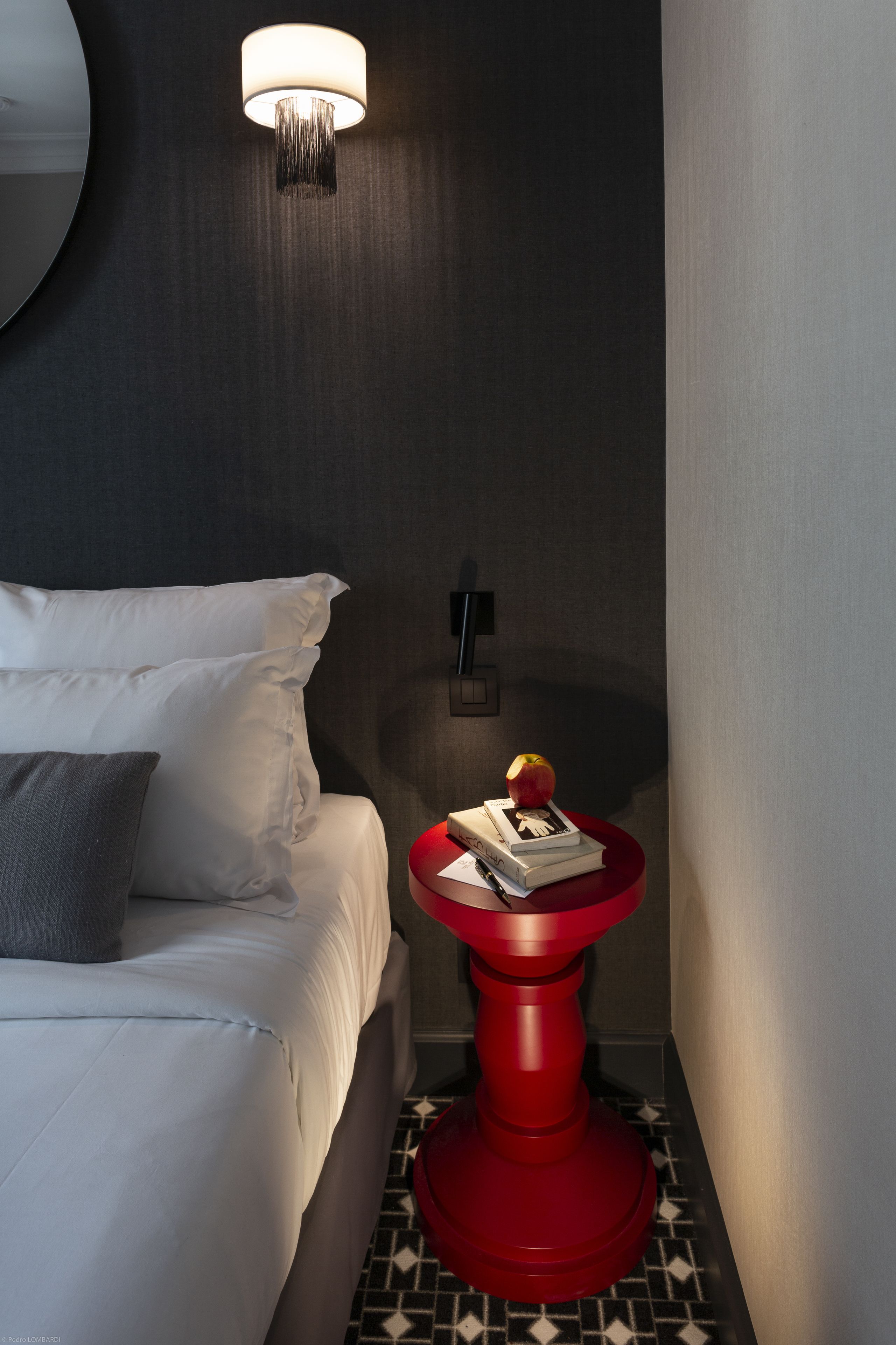 A hotel room with a bed, a red side table with books and an apple, a wall-mounted light, and a patterned floor.