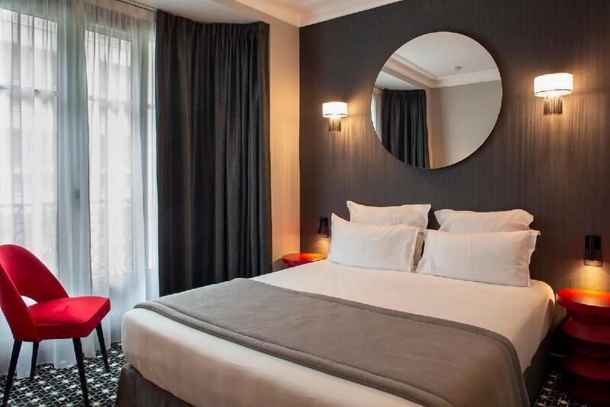 A hotel room with a bed, a red chair, a round mirror, and two wall-mounted lights.