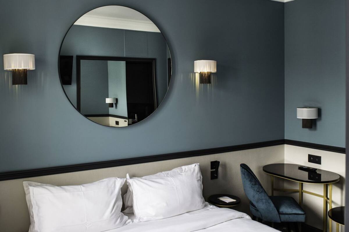 A hotel room with a bed, a round mirror, two wall-mounted lamps, a small round table, a blue chair, and a nightstand.