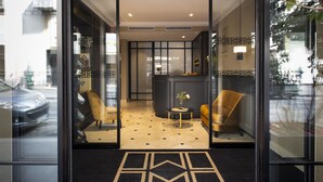 Un vestíbulo de hotel con un suelo de alfombra a blanco y negro, un mostrador de recepción y una zona de estar con un sillón amarillo.