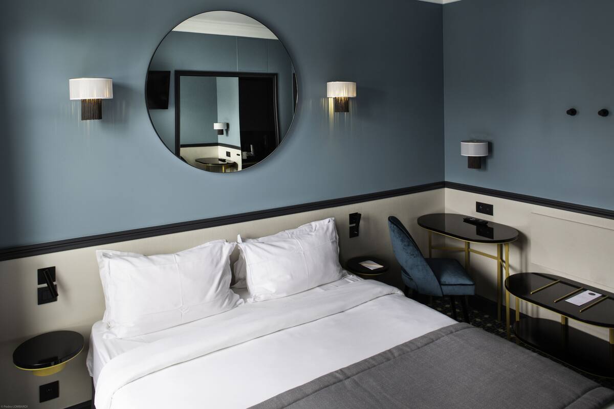 A hotel room with a bed, a round mirror, two wall-mounted lamps, a small round table, and a blue chair.