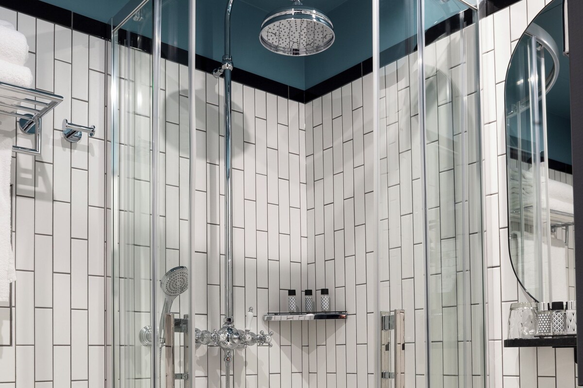 Un baño moderno con ducha envuelta en vidrio, paredes cubiertas de azulejos blancos, un lavamanos blanco y un espejo sobre él.