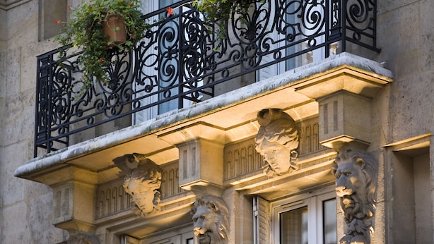 Fachada de edificio con barandillas de hierro ornamentadas, esculturas de leones de piedra y una planta en maceta.