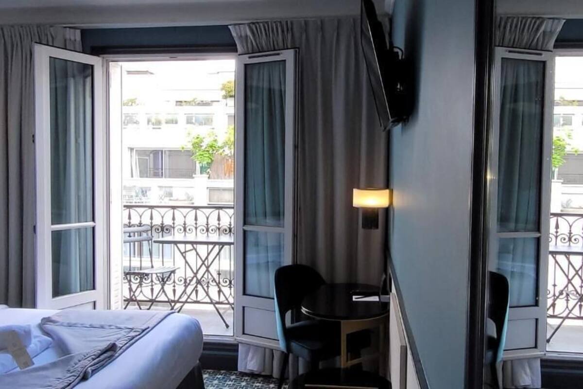 A hotel room with a bed, a desk, and a balcony with a view of buildings and a fence.