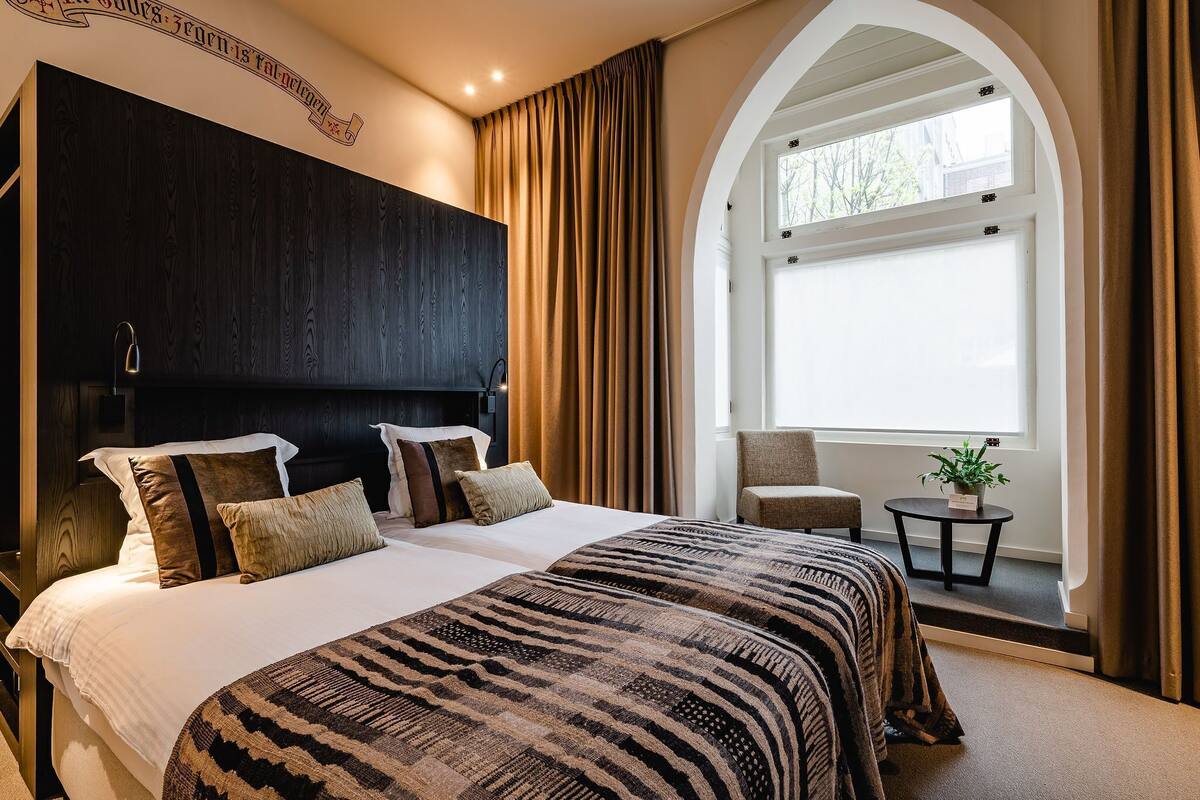 A bedroom with a large bed, a dark headboard, a window with curtains, a small table with a plant, and a chair.