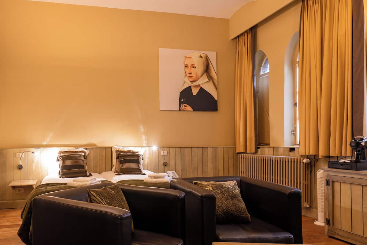 A hotel room with a bed, two chairs, a painting on the wall, a radiator, and a window with curtains.