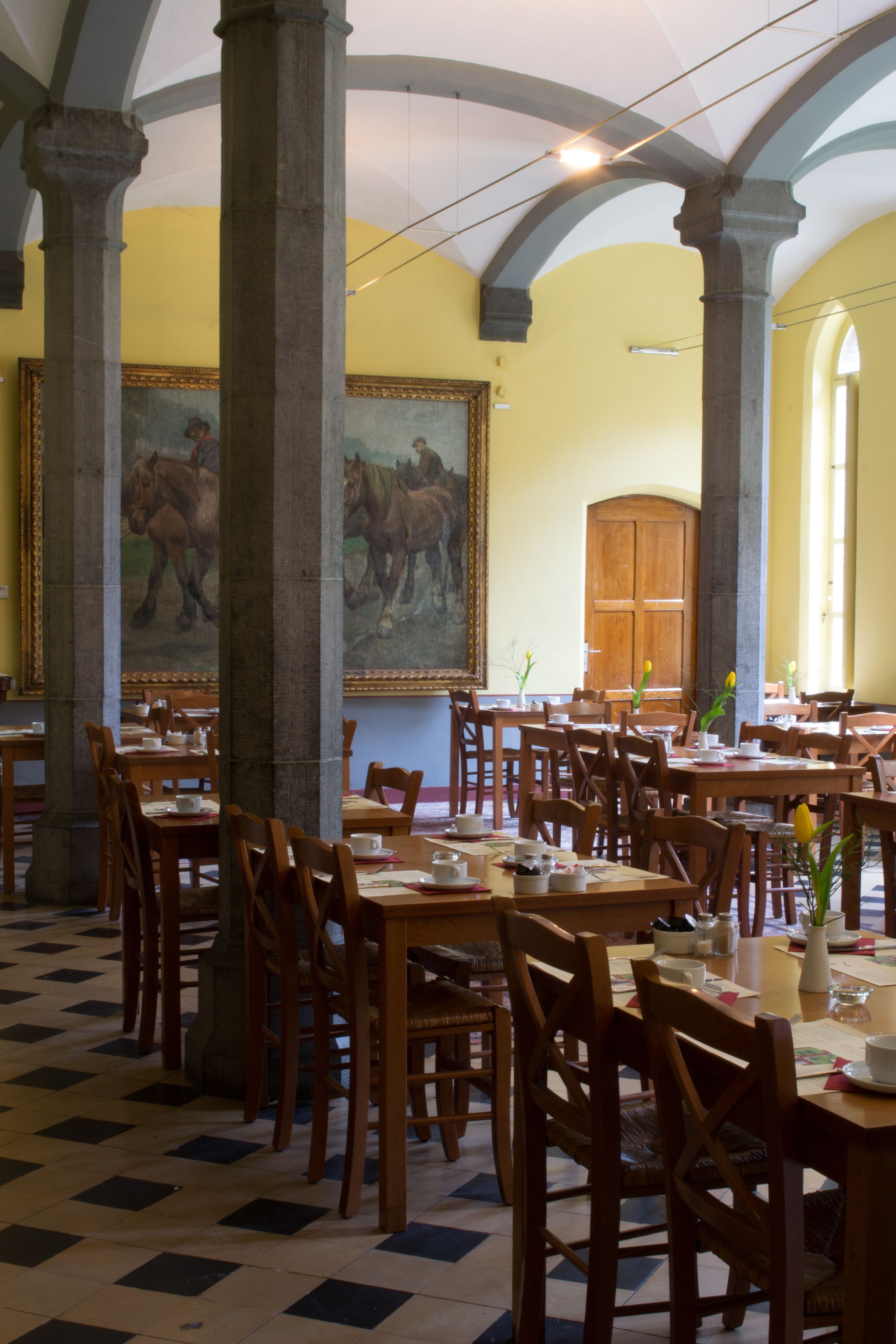 A dining hall with wooden tables and chairs, large columns, and a mural on the wall.