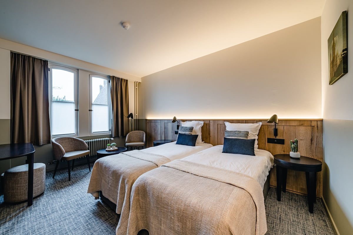 A hotel room with two beds, a chair, a small table, and a window with curtains.