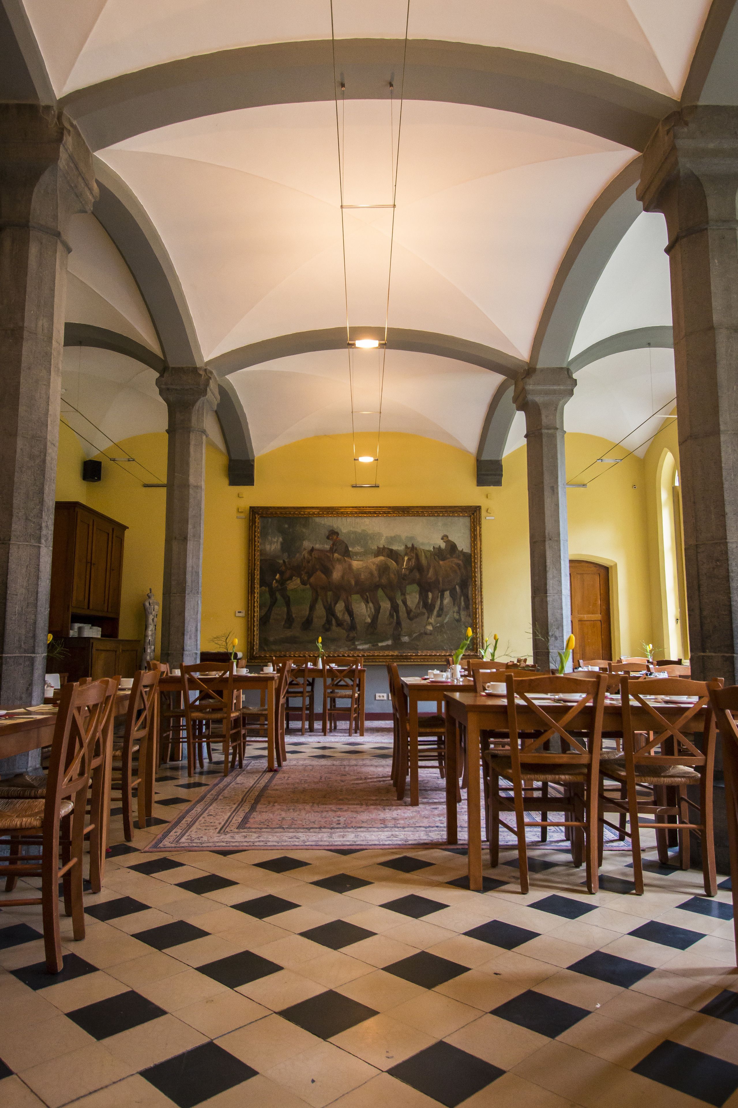 Een eetzaal met hoge plafonds, schachoottegoud, houten tafels en stoelen, en een groot schilderij aan de muur.