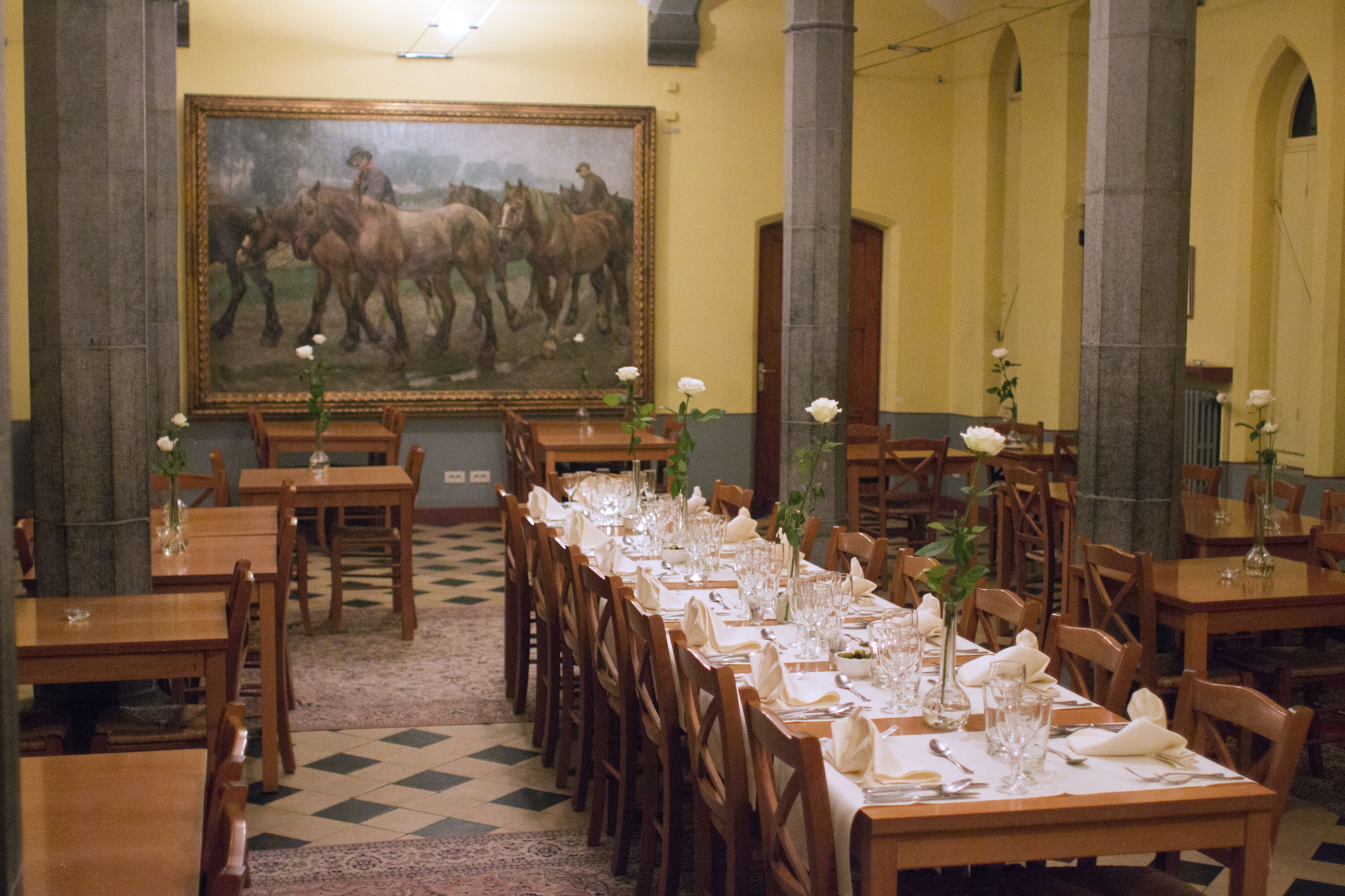 Une salle à manger avec de longues tables dressées pour le repas, des chaises en bois et un grand tableau accroché au mur.