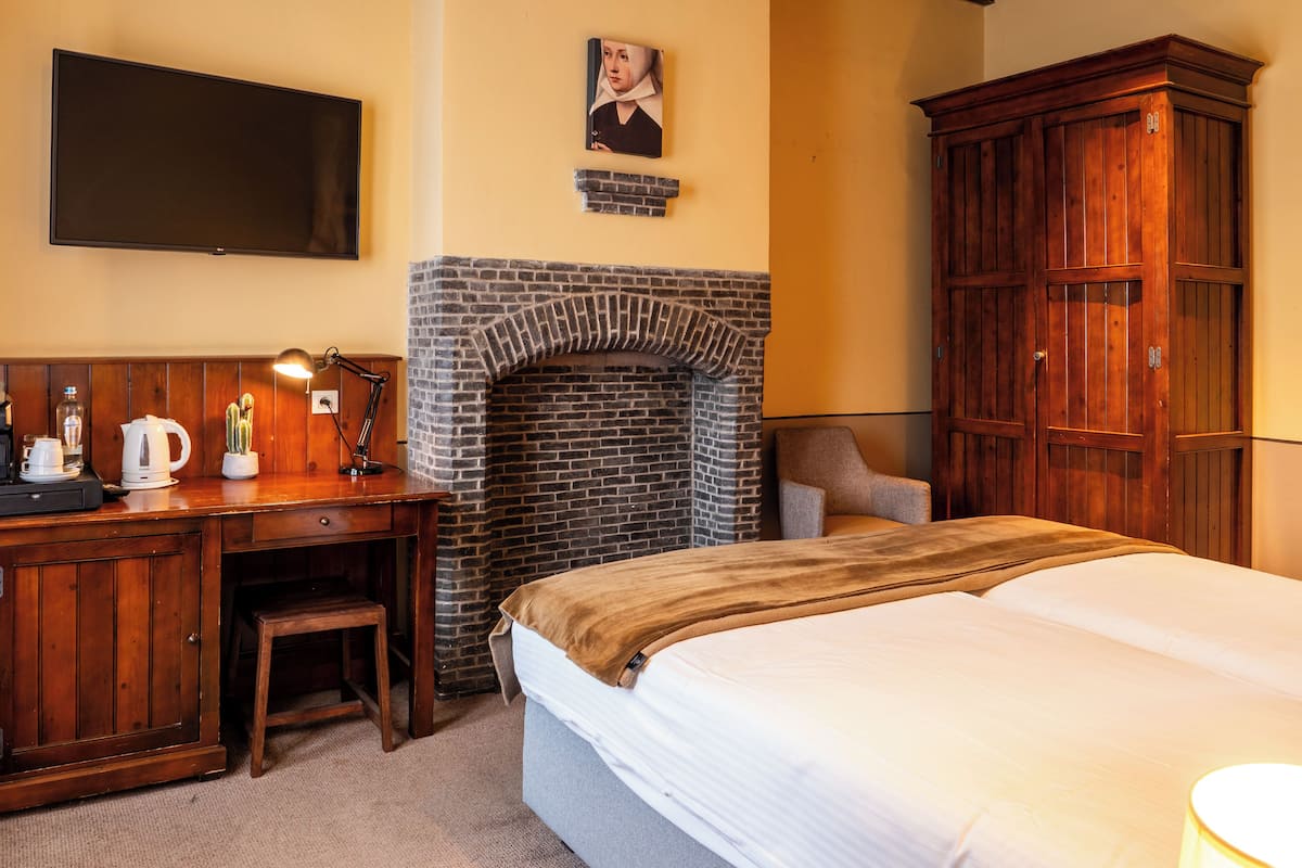 A bedroom with a brick fireplace, a bed, a desk with a chair, a television, and wooden cabinets.