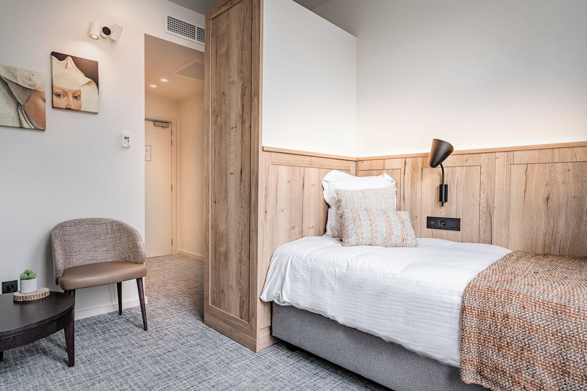 A bedroom with a bed, a chair, a small table, and a wall-mounted lamp.