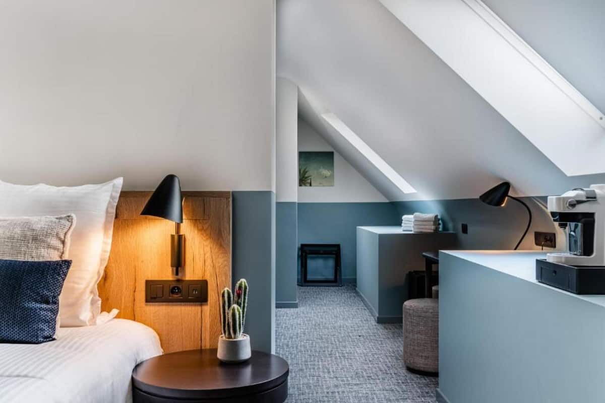 A modern hotel room with a bed, bedside table, lamp, and a small potted plant. There is a kitchenette area with a coffee machine and a counter.