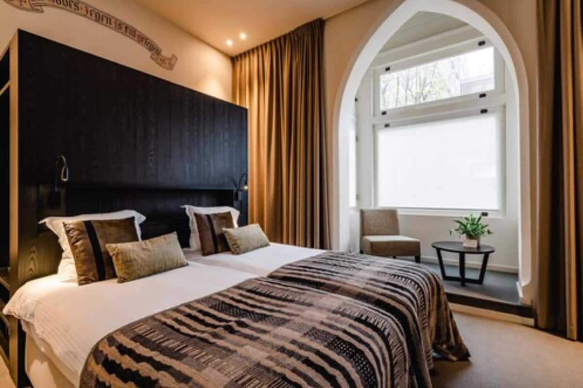 A hotel room with a large bed, a dark headboard, a small table with a plant, and a window with curtains.