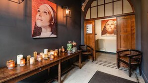 A cozy interior with a wooden bench lined with candles, a large portrait on the wall, and a rustic wooden door. - Monasterium PoortAckere (Ghent)
