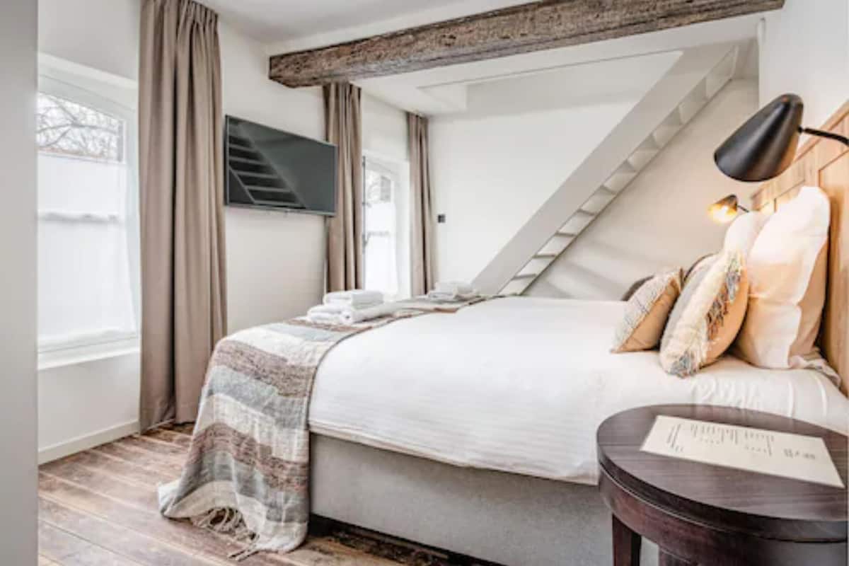 A bedroom with a bed, a TV mounted on the wall, a staircase, and a small round table with a lamp.