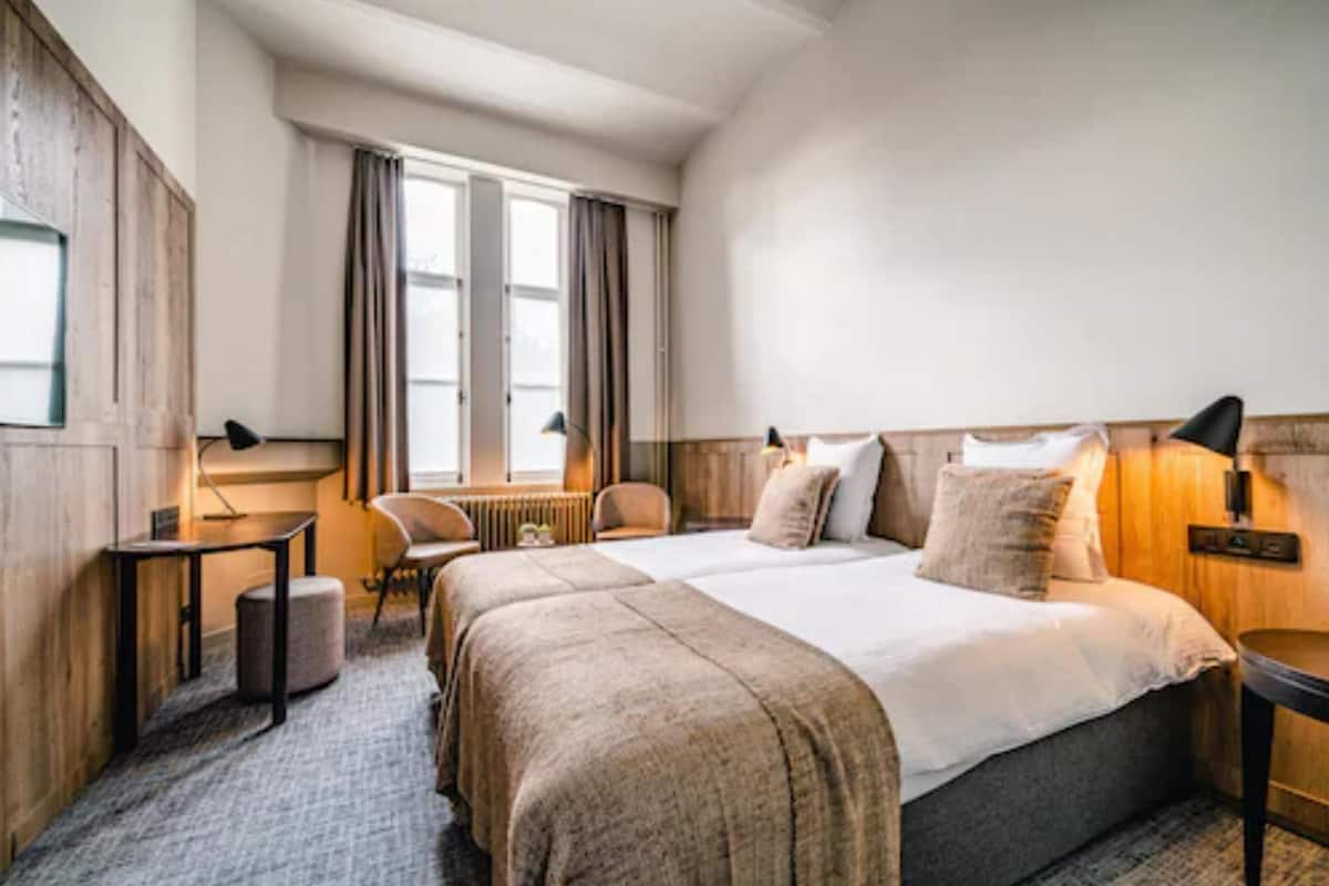 A hotel room with a large bed, two bedside tables, a chair, a small table, and a window with curtains.