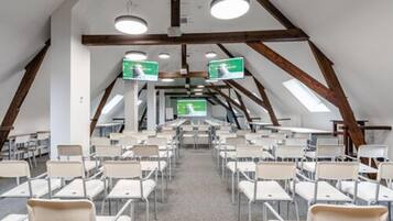 A conference room with white chairs arranged in rows, a high ceiling with wooden beams, and two screens at the front.