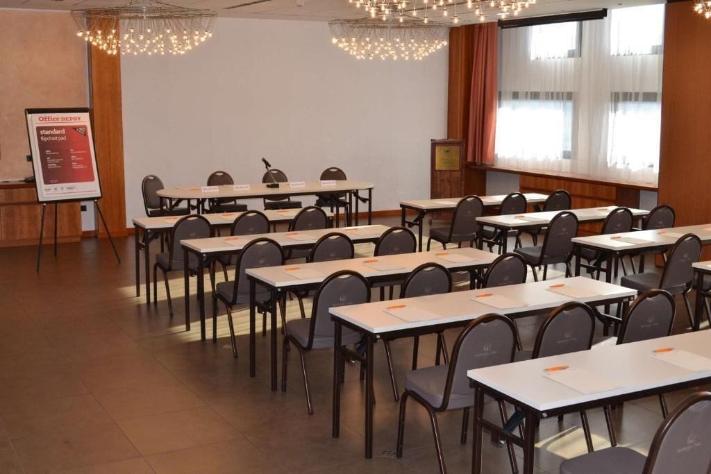 Une salle de conférence équipée de plusieurs tables et chaises, d’un pupitre et d’une grande fenêtre avec des rideaux.