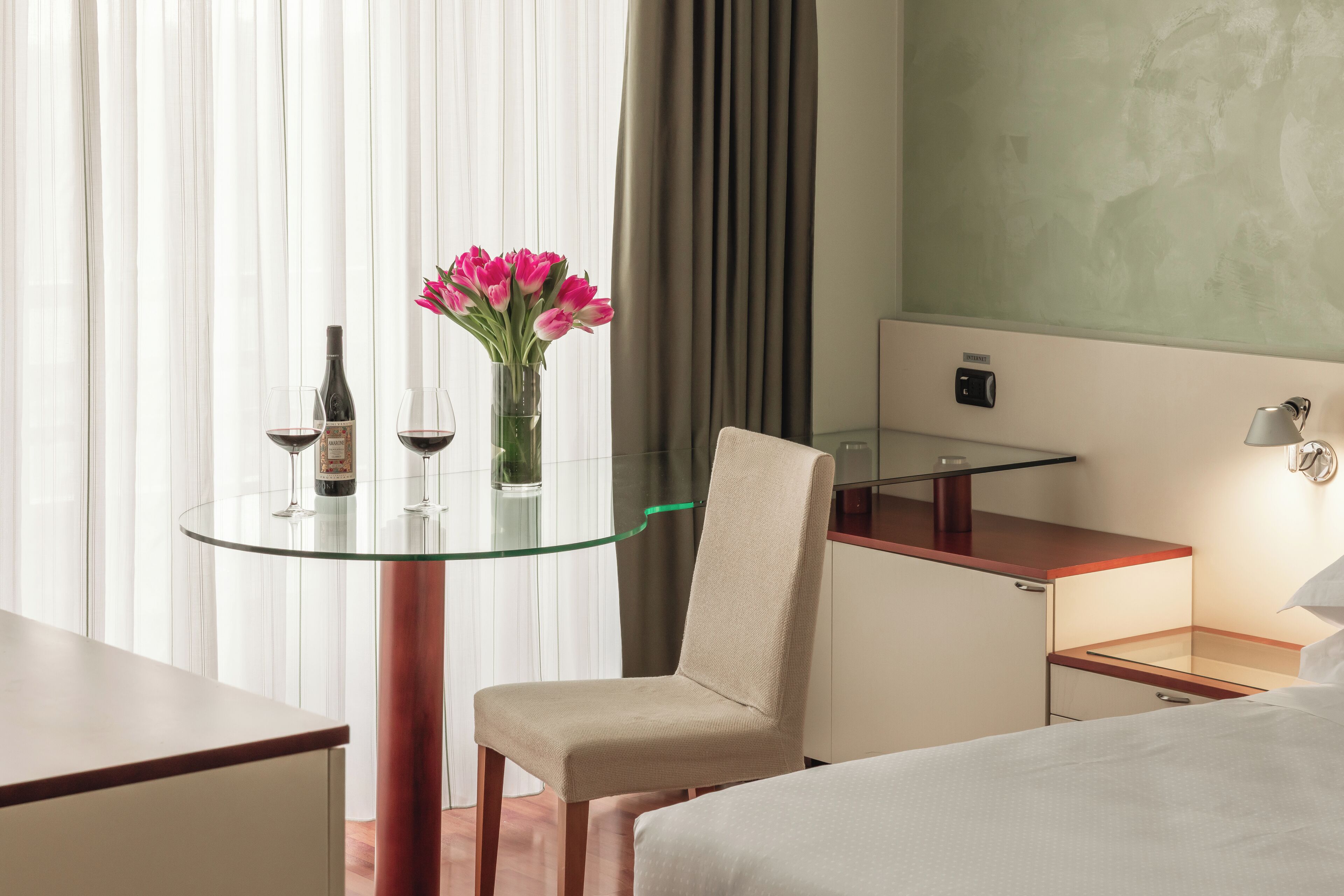 A hotel room with a glass table, a chair, a bed, a desk, and a nightstand.