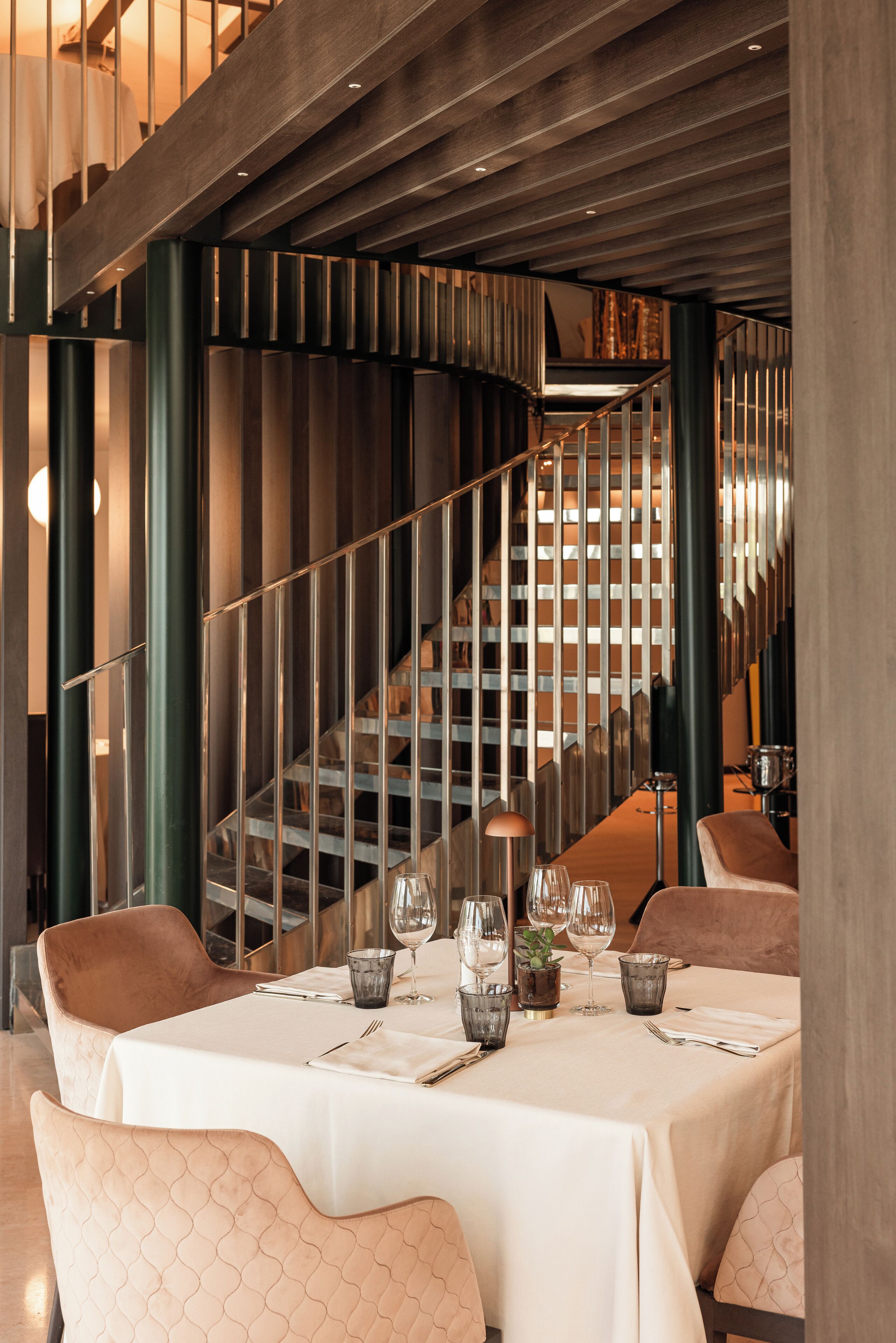 Ein Restaurant mit modernem Ambiente, das eine Wendeltreppe, Essplätze mit Gläsern und Besteck sowie gepolsterte Stühle zeigt.