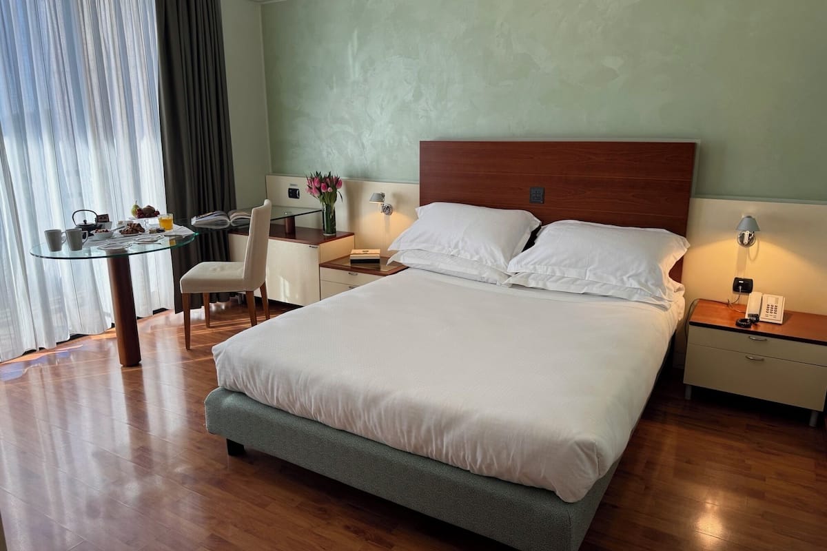 A hotel room with a large bed, a desk with a chair, a bedside table with a lamp, and a window with curtains.