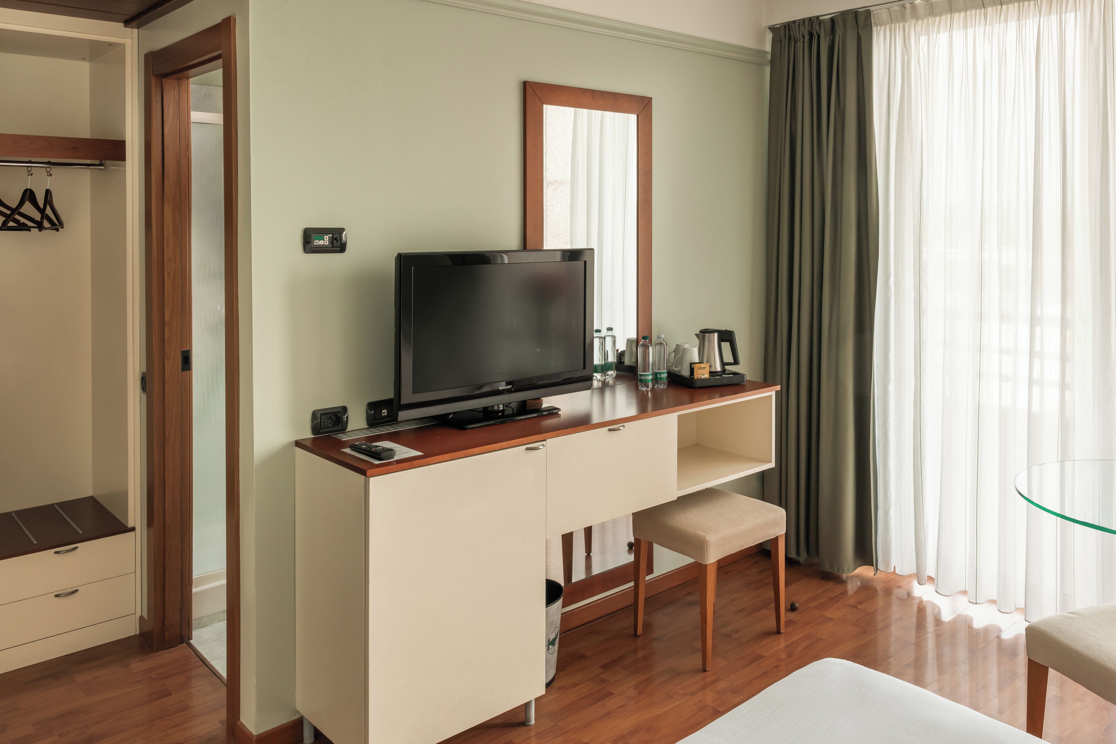 Una habitación de hotel con un escritorio de madera, un televisor, un pequeño banco y una mesa de vidrio.