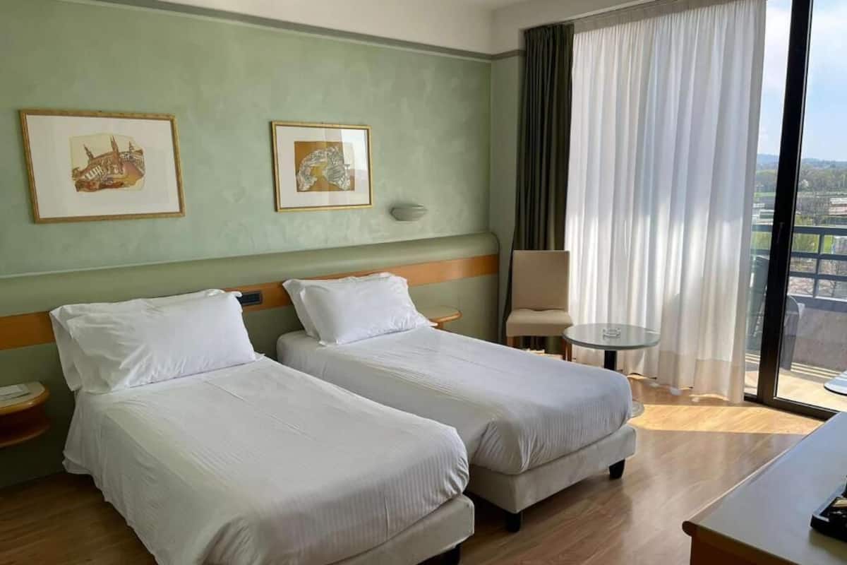 A hotel room with two single beds, a small table, a chair, and a large window with curtains.