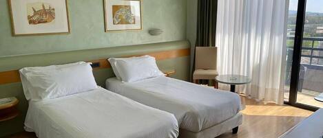 Habitación de hotel con dos camas individuales, una mesita pequeña, una silla y un ventanal grande con cortinas.