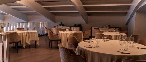 Un restaurant avec des tables rondes, des chaises rembourrées et un plafond haut avec des poutres apparentes.