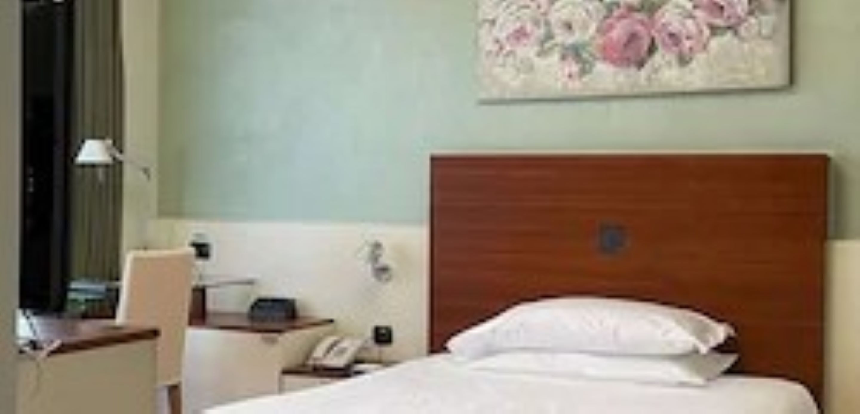 A hotel room with a wooden headboard, a bed with white linens, a desk with a lamp, and a floral painting on the wall.