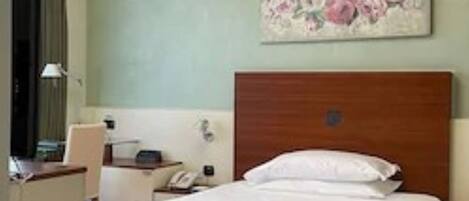 A hotel room with a wooden headboard, a bed with white linens, a desk with a lamp, and a floral painting on the wall.