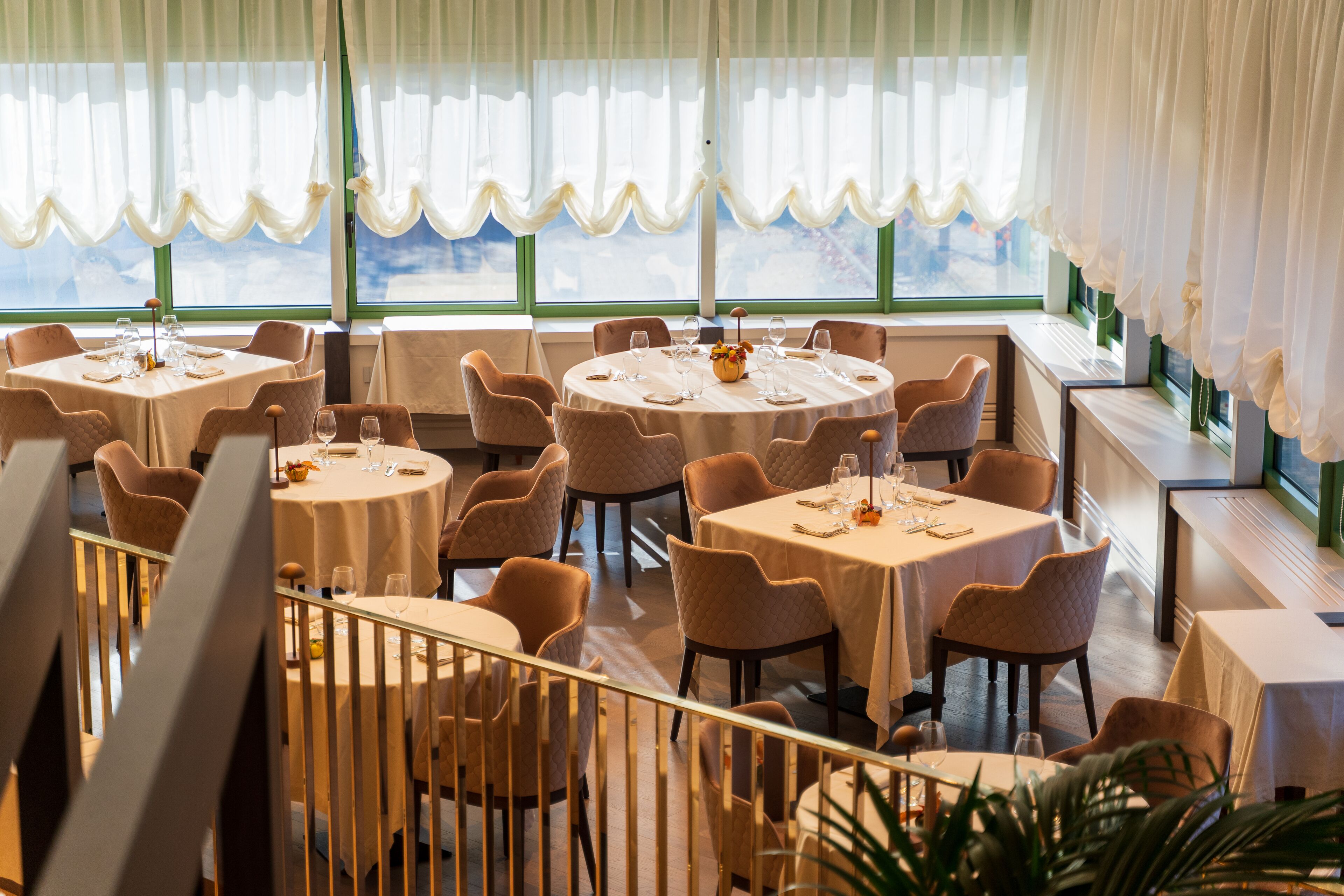A restaurant with round tables and chairs, large windows with curtains, and a view of the outdoors.